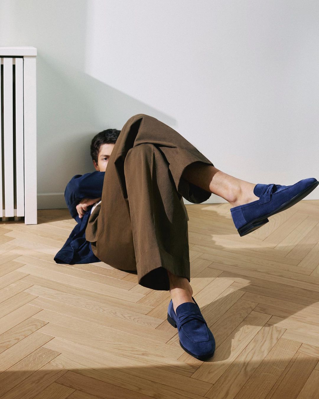 Stylish indoor shot of a man lying on a wooden floor, showcasing trendy brown trousers and blue suede loafers, illuminated by natural light.