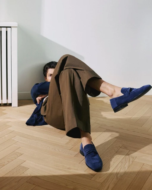 Stylish indoor shot of a man lying on a wooden floor, showcasing trendy brown trousers and blue suede loafers, illuminated by natural light.