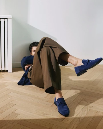 Stylish indoor shot of a man lying on a wooden floor, showcasing trendy brown trousers and blue suede loafers, illuminated by natural light.