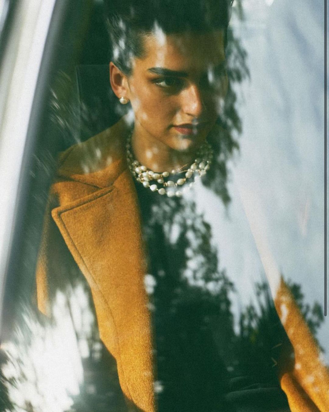 Portrait of a woman with dark hair and a pearl necklace, wearing a tan coat, gazing through a window with tree reflections.