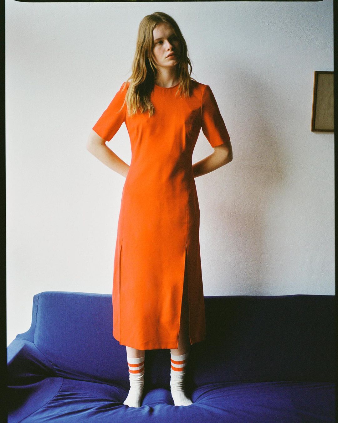 Woman in an orange dress and white socks standing behind a dark blue couch against a white wall.