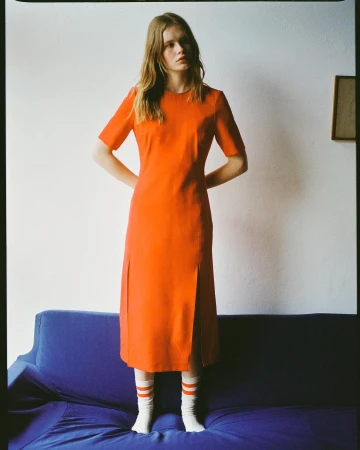 Woman in an orange dress and white socks standing behind a dark blue couch against a white wall.
