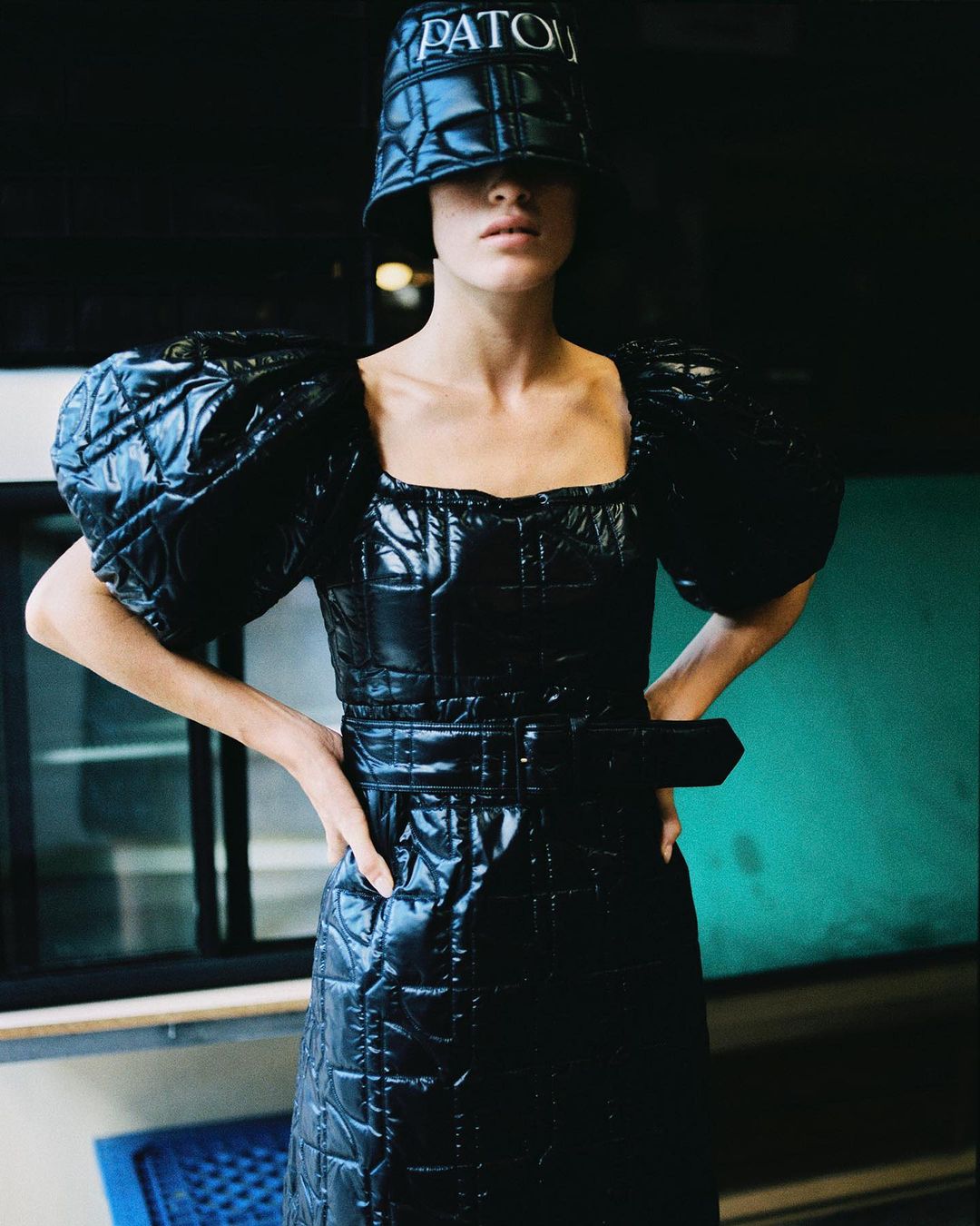 Woman poses in a shiny, quilted black puffer dress and bucket hat with 'PATOU' logo, standing indoors with hands on her hips.
