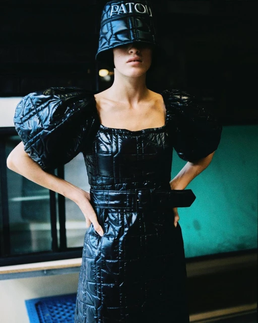 Woman poses in a shiny, quilted black puffer dress and bucket hat with 'PATOU' logo, standing indoors with hands on her hips.