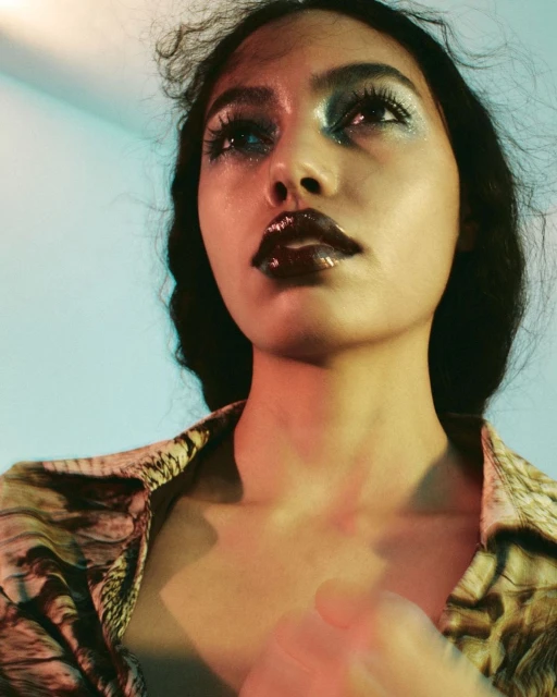Portrait of a stylish young woman with elaborate makeup and a patterned shirt, looking up against a softly colored background.