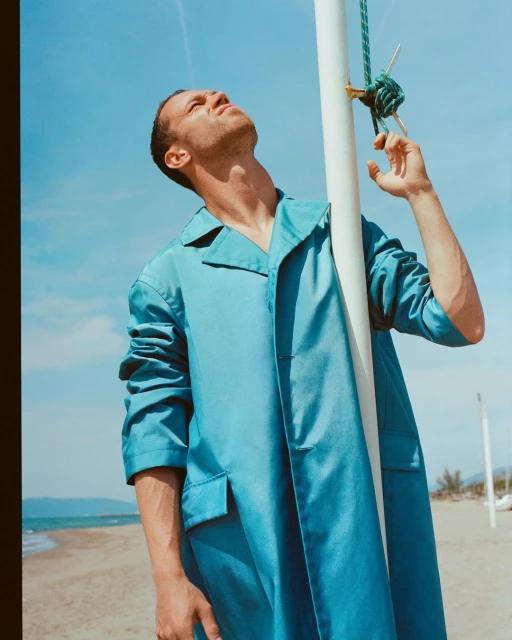 Man in a turquoise jacket stands on a beach, looking upward toward a white pole with a rope knot under a clear blue sky.