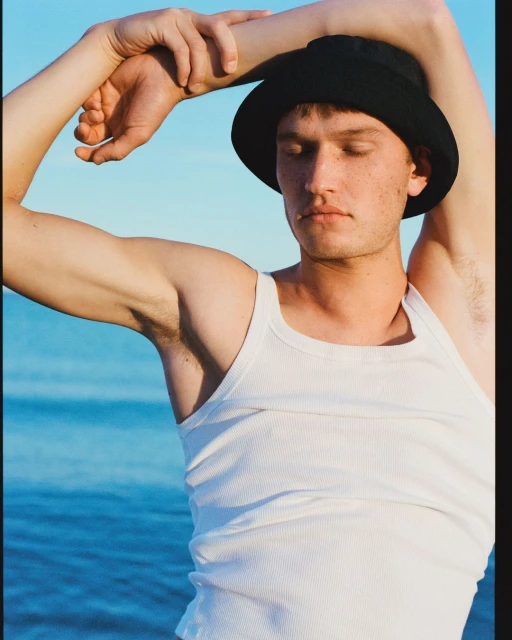 Portrait of a young adult man with closed eyes wearing a black hat and white tank top, posing outdoors by a blue sea and clear sky.