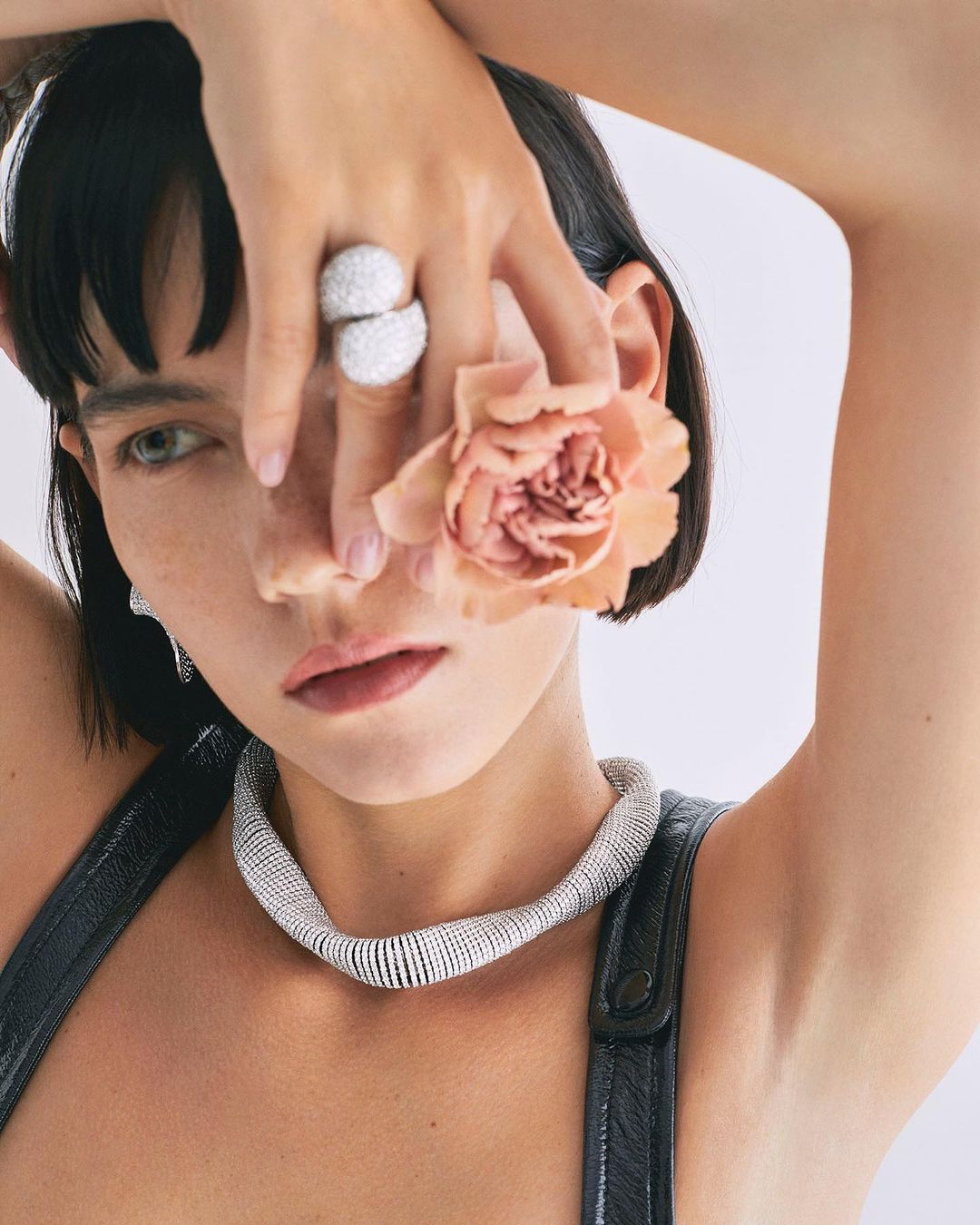 Brunette woman poses indoors, wearing a metallic necklace and holding a peach flower to her face, accessorized with a large silver ring.