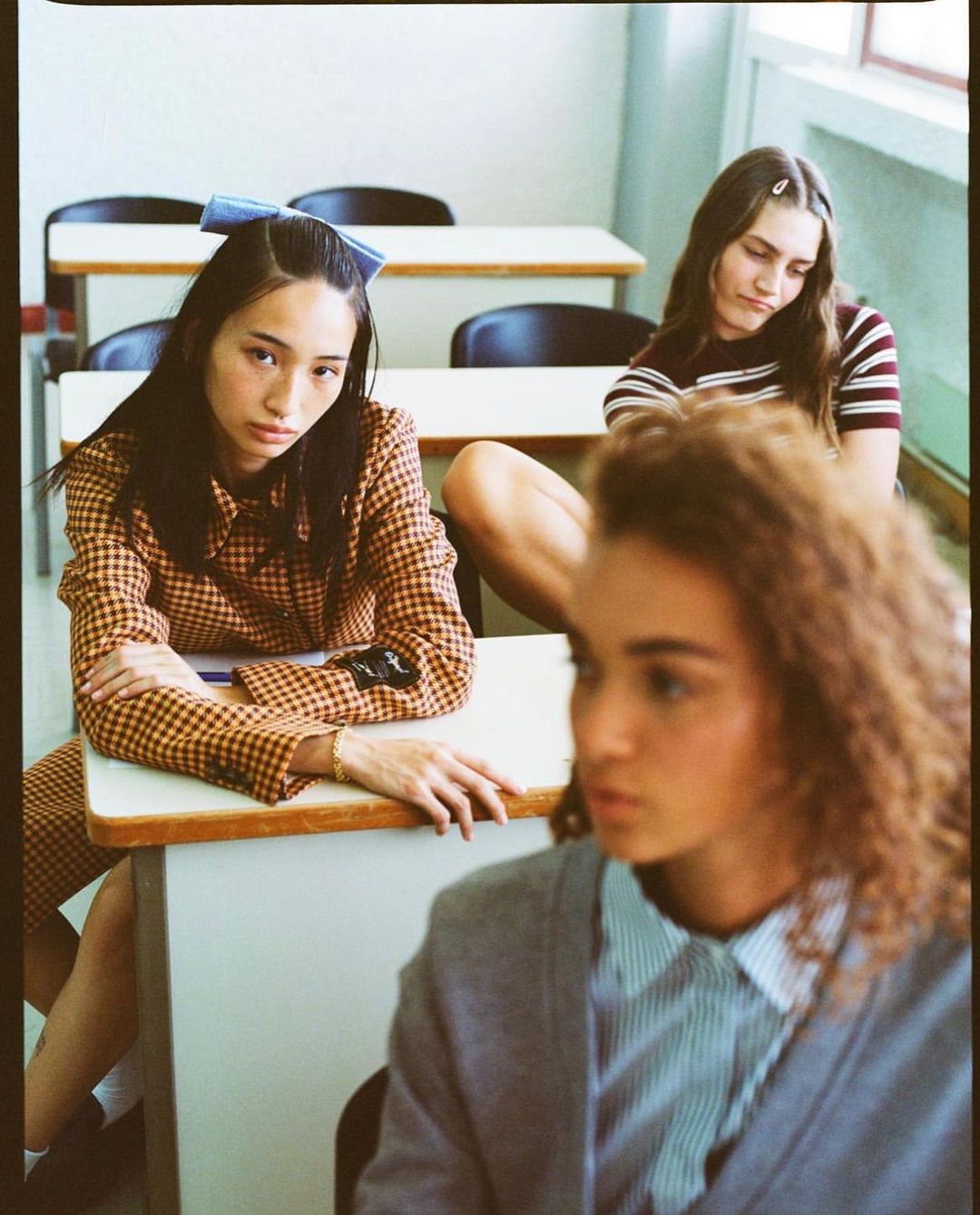 Three young women in an indoor classroom setting, wearing casual attire and posing at desks. One wears a blue bow. School concept.