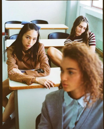 Three young women in an indoor classroom setting, wearing casual attire and posing at desks. One wears a blue bow. School concept.