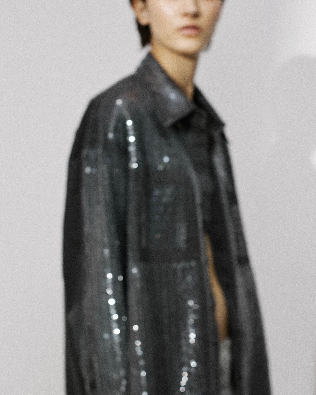 Fashion portrait of a young adult woman wearing a sequined jacket indoors against a white wall, shot in soft studio lighting.