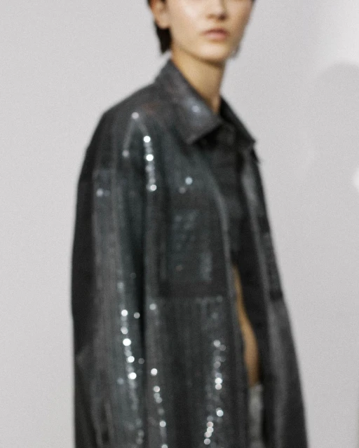 Fashion portrait of a young adult woman wearing a sequined jacket indoors against a white wall, shot in soft studio lighting.
