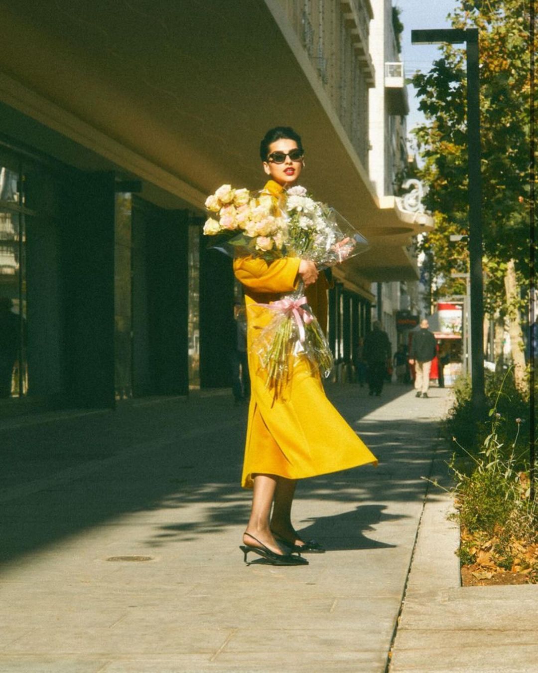 A woman in a bright yellow dress holds a large bouquet of white and pink flowers on a sunny city street with buildings behind her.