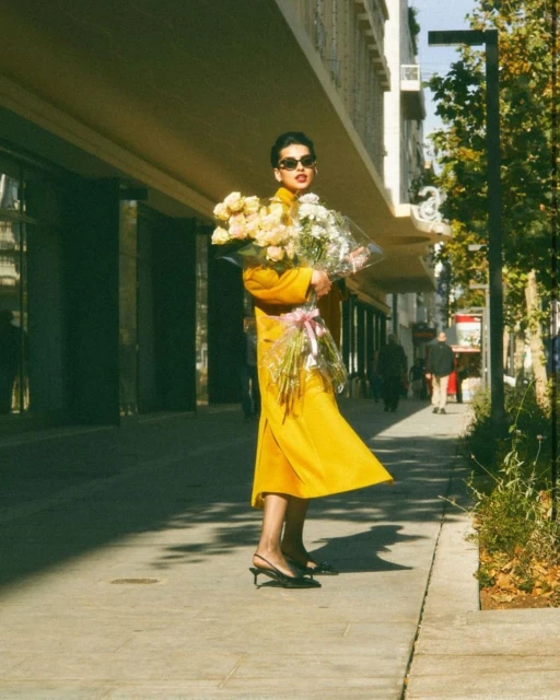 A woman in a bright yellow dress holds a large bouquet of white and pink flowers on a sunny city street with buildings behind her.
