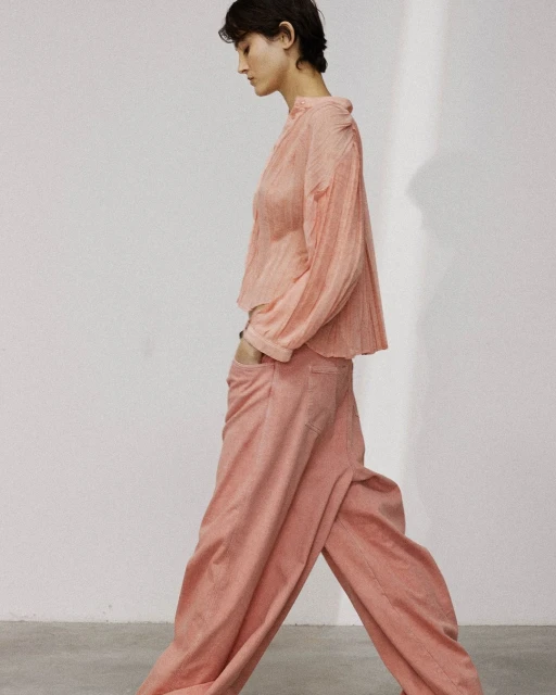 An adult female walking in a light salmon colored, long sleeve blouse and matching pants against a plain, neutral background in studio lighting.