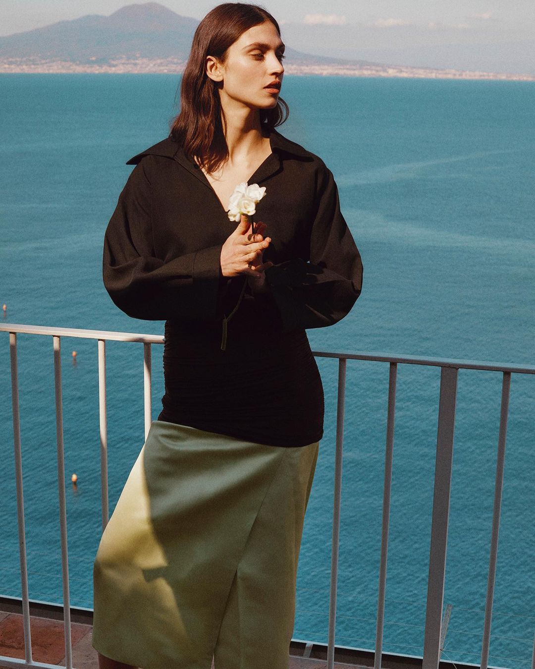 A young adult female model poses outdoors by a railing, wearing a black shirt and light green skirt, with a distant sea backdrop.