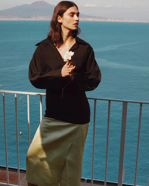 A young adult female model poses outdoors by a railing, wearing a black shirt and light green skirt, with a distant sea backdrop.