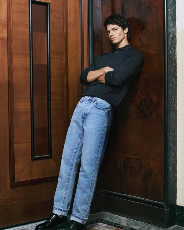 Young male model leaning against a wooden door wearing a dark polo shirt and light blue denim jeans