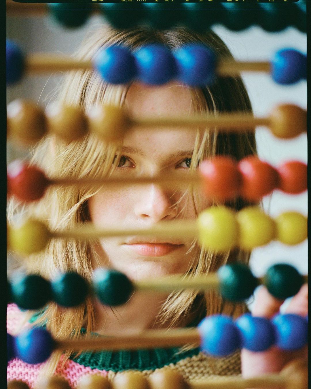 Portrait of a young woman looking through a vintage abacus with colorful beads in a soft, filtered light, and wearing a pastel sweater.