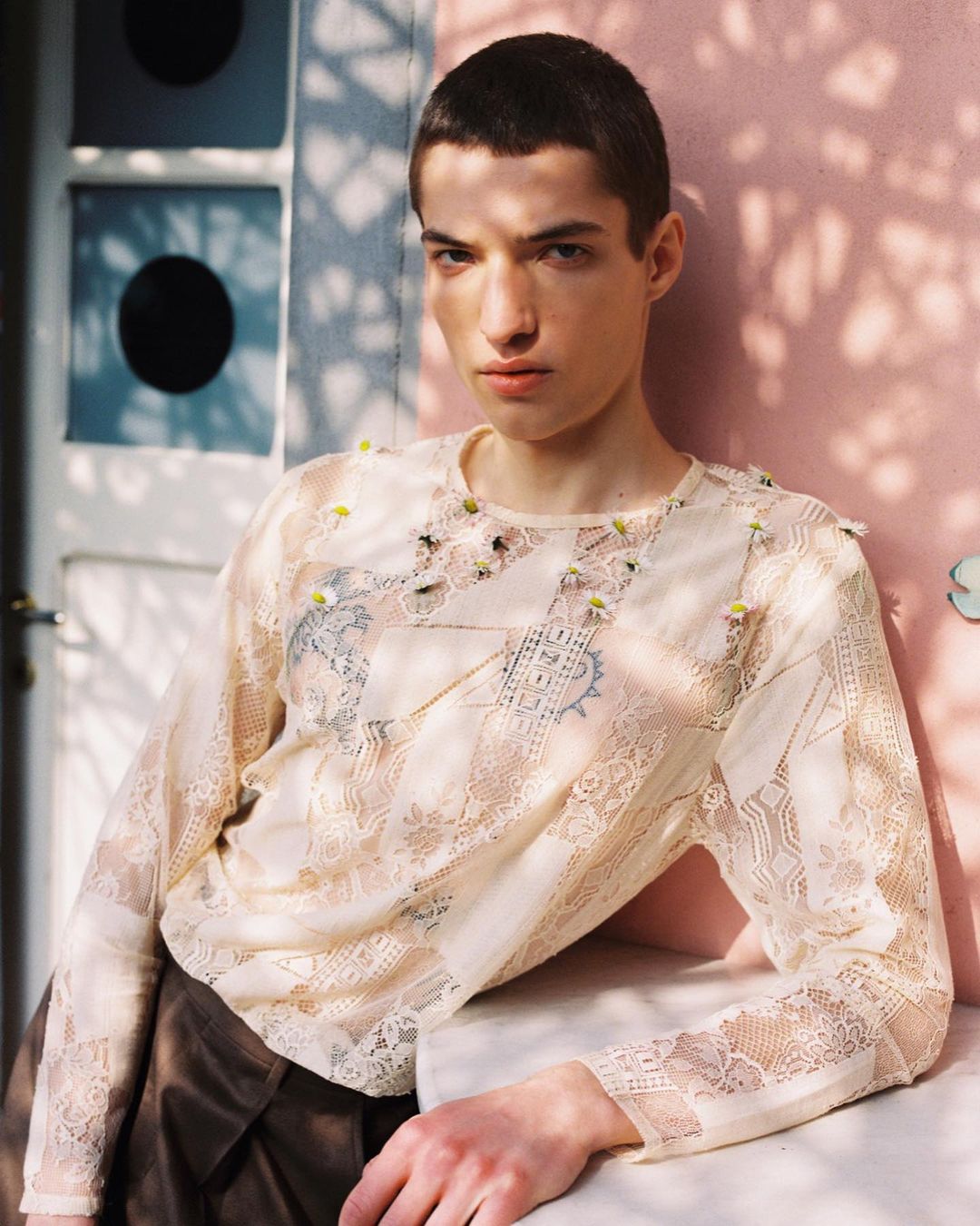 Portrait of a young adult male wearing a lace shirt, leaning against a white surface with a pink wall and window in the background.