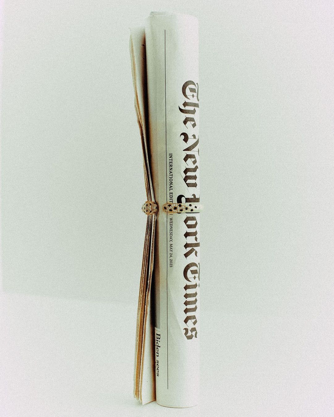 Rolled-up New York Times newspaper, secured with a decorative ring, set against a plain background.