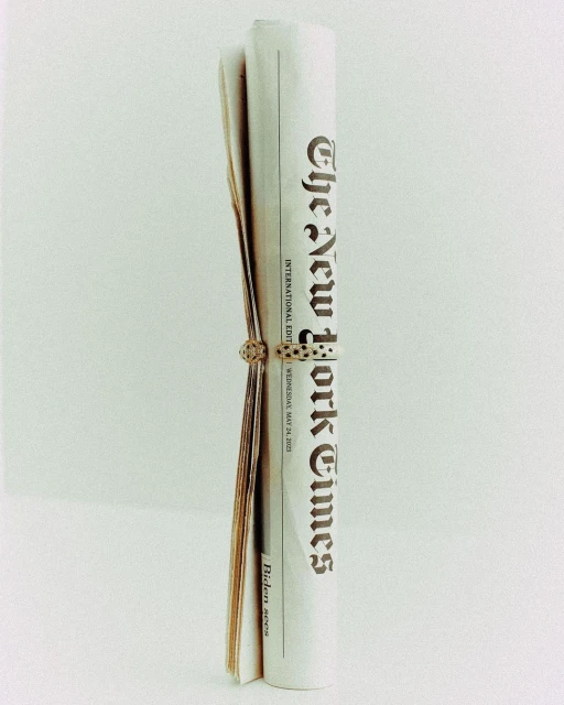 Rolled-up New York Times newspaper, secured with a decorative ring, set against a plain background.