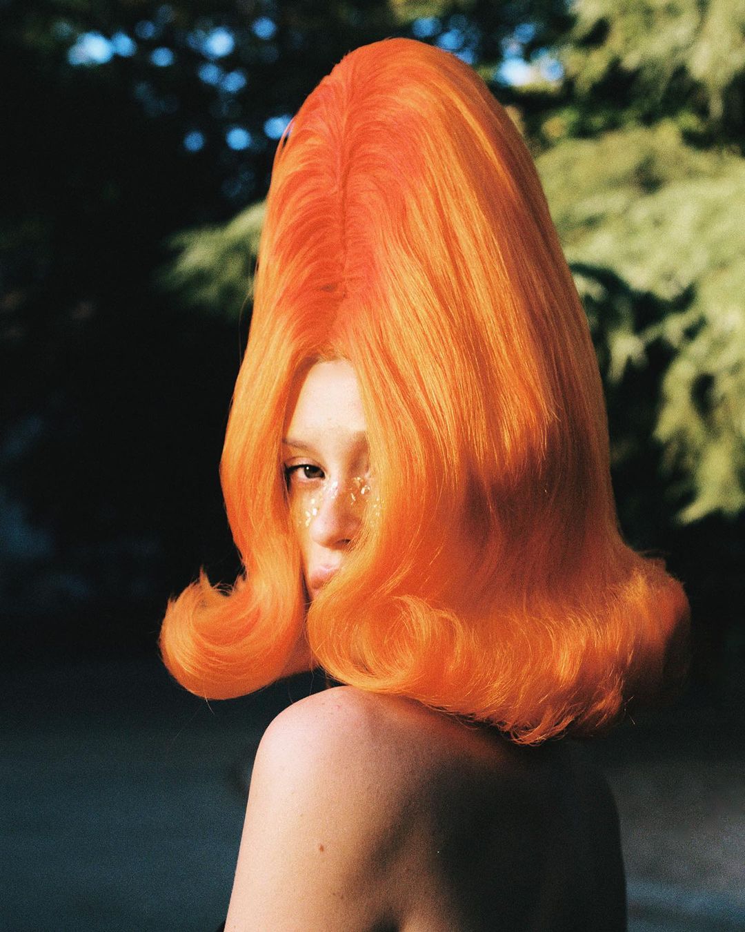 Woman in vibrant orange beehive wig poses outdoors with glitter makeup, bare shoulders, and blurred green foliage background.