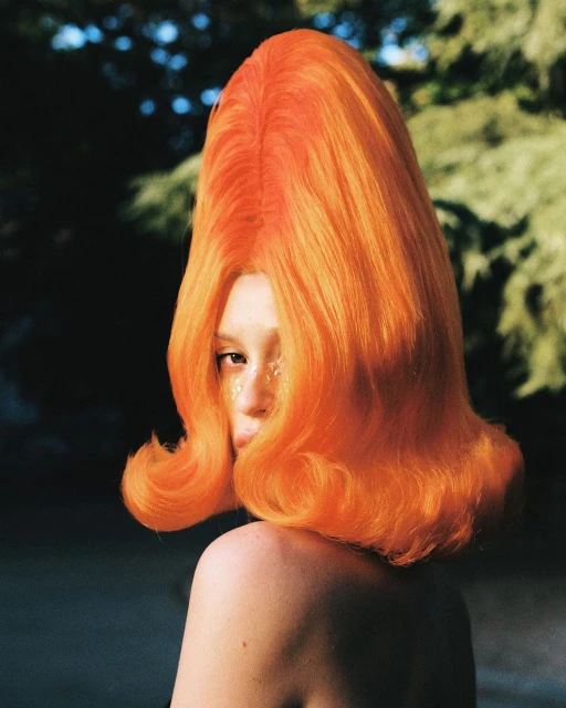 Woman in vibrant orange beehive wig poses outdoors with glitter makeup, bare shoulders, and blurred green foliage background.