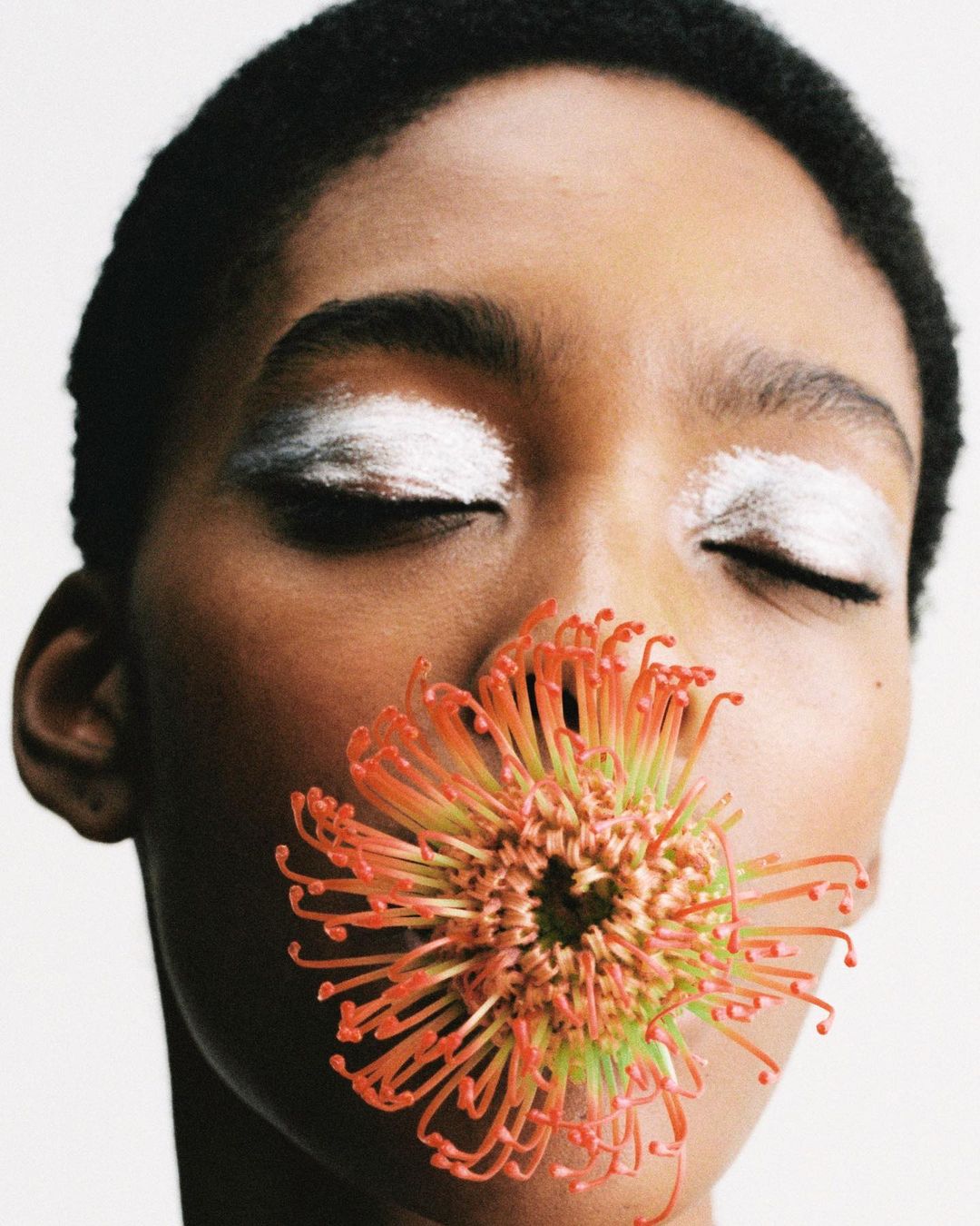 Close-up portrait of a woman with closed eyes, wearing white eye makeup, and holding an orange protea flower near her mouth in a bright studio.