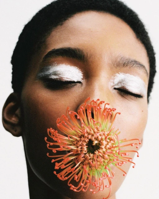 Close-up portrait of a woman with closed eyes, wearing white eye makeup, and holding an orange protea flower near her mouth in a bright studio.