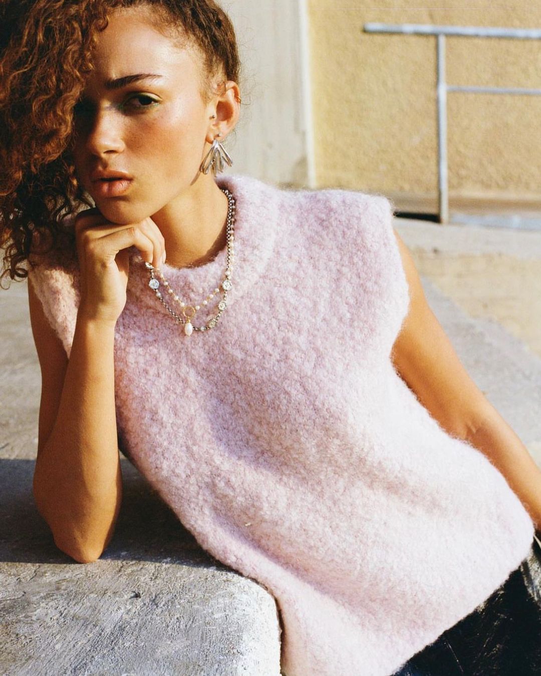 Model with curly hair wearing a pale pink knit vest and layered necklaces leaning on a light stone ledge outdoors