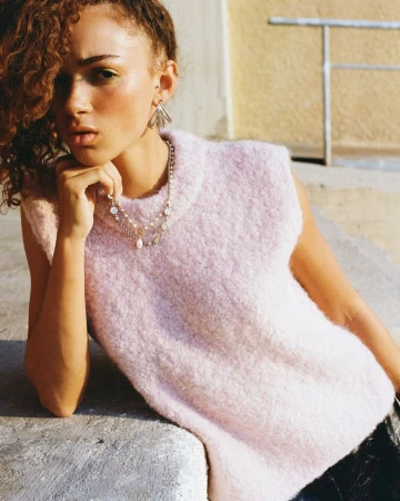 Model with curly hair wearing a pale pink knit vest and layered necklaces leaning on a light stone ledge outdoors