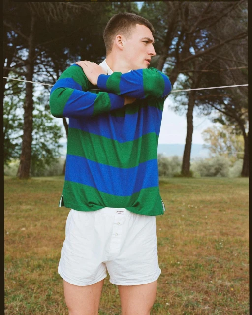 Athletic man in striped polo shirt and white shorts standing in grassy clearing surrounded by trees on a sunny day in the countryside.