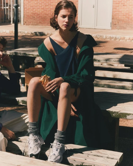 A young adult woman sits outside on a wooden bench in a blue dress and green jacket, holding a sandwich, with two other people.
