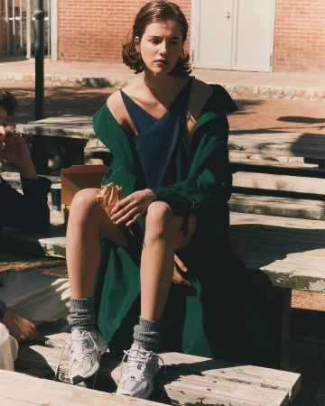 A young adult woman sits outside on a wooden bench in a blue dress and green jacket, holding a sandwich, with two other people.