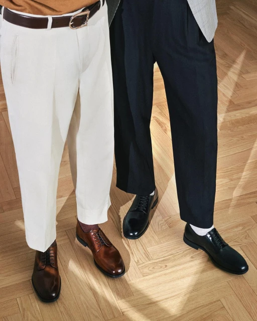 Two men showcase fashion, one in white trousers and brown shoes, the other in dark trousers and black shoes, set on a herringbone wood floor.