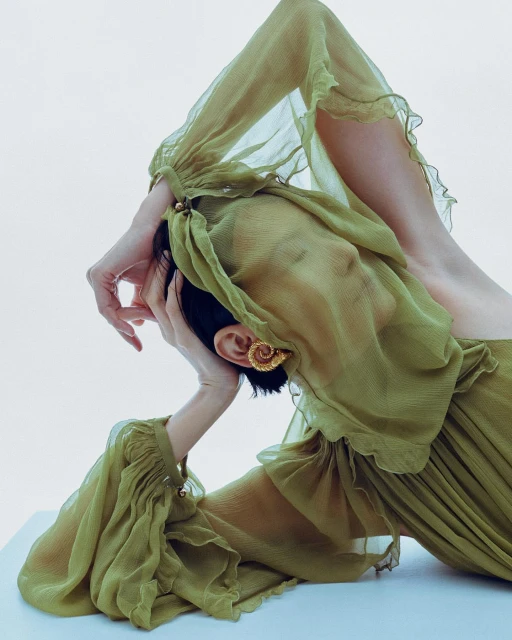 A woman in a flowing, light green dress poses in a studio, creating an editorial fashion image that highlights the dress's texture and color.