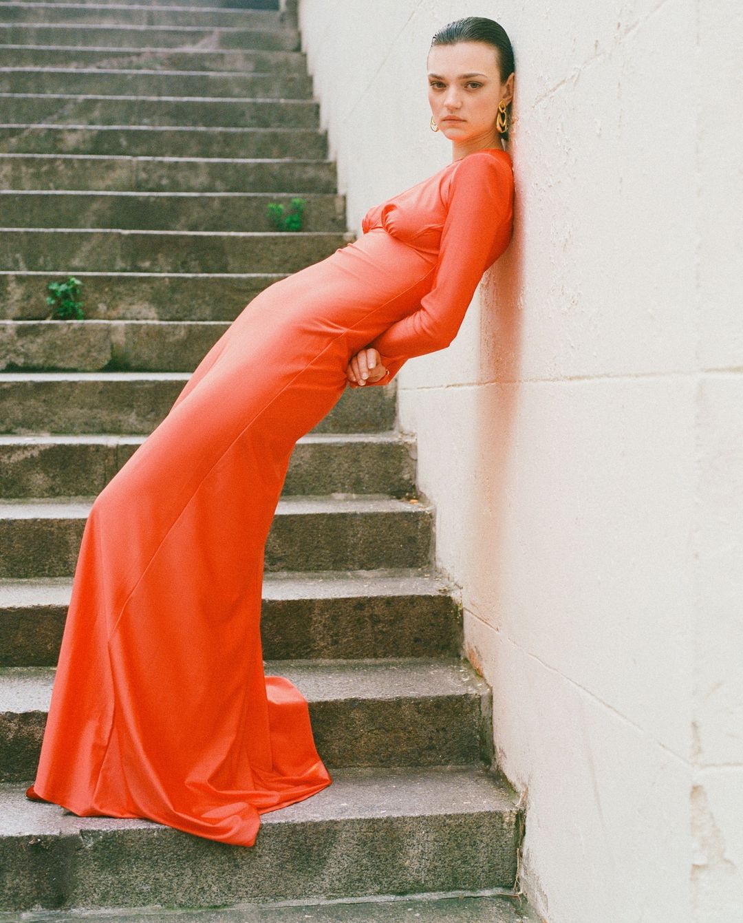 Model in long orange dress leaning against a concrete wall beside stone stairs, captured by Alessandro Burzigotti.