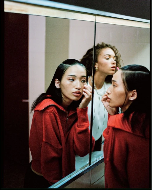 Two women pose in a bathroom with an Asian woman reflected in a mirror wearing a red knit jacket and neutral makeup, looking at her reflection.
