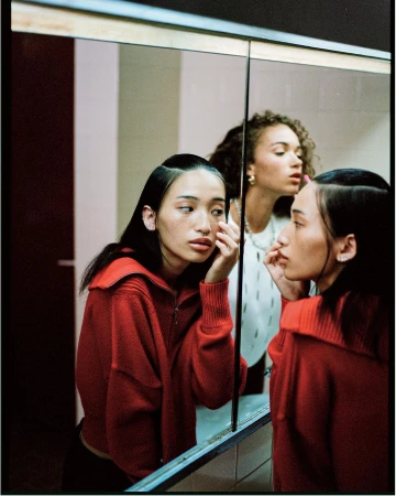 Two women pose in a bathroom with an Asian woman reflected in a mirror wearing a red knit jacket and neutral makeup, looking at her reflection.