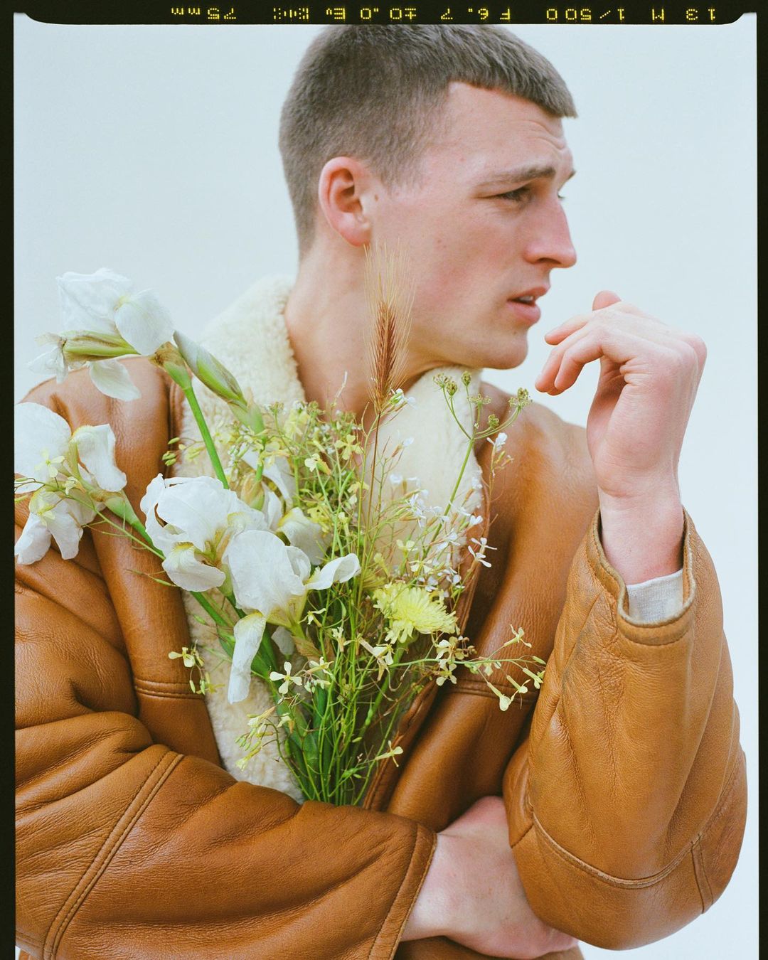 Portrait of a young man with short brown hair in a brown shearling jacket, holding a bouquet of white and yellow flowers indoors.