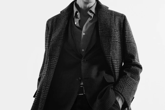 Man wearing a tweed coat, patterned button-up shirt, and suit jacket against a plain light background.