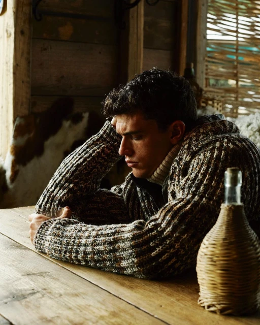 Indoor portrait of a thoughtful young adult male sitting at a wooden table, wearing a patterned knit sweater, with a woven bottle prop.