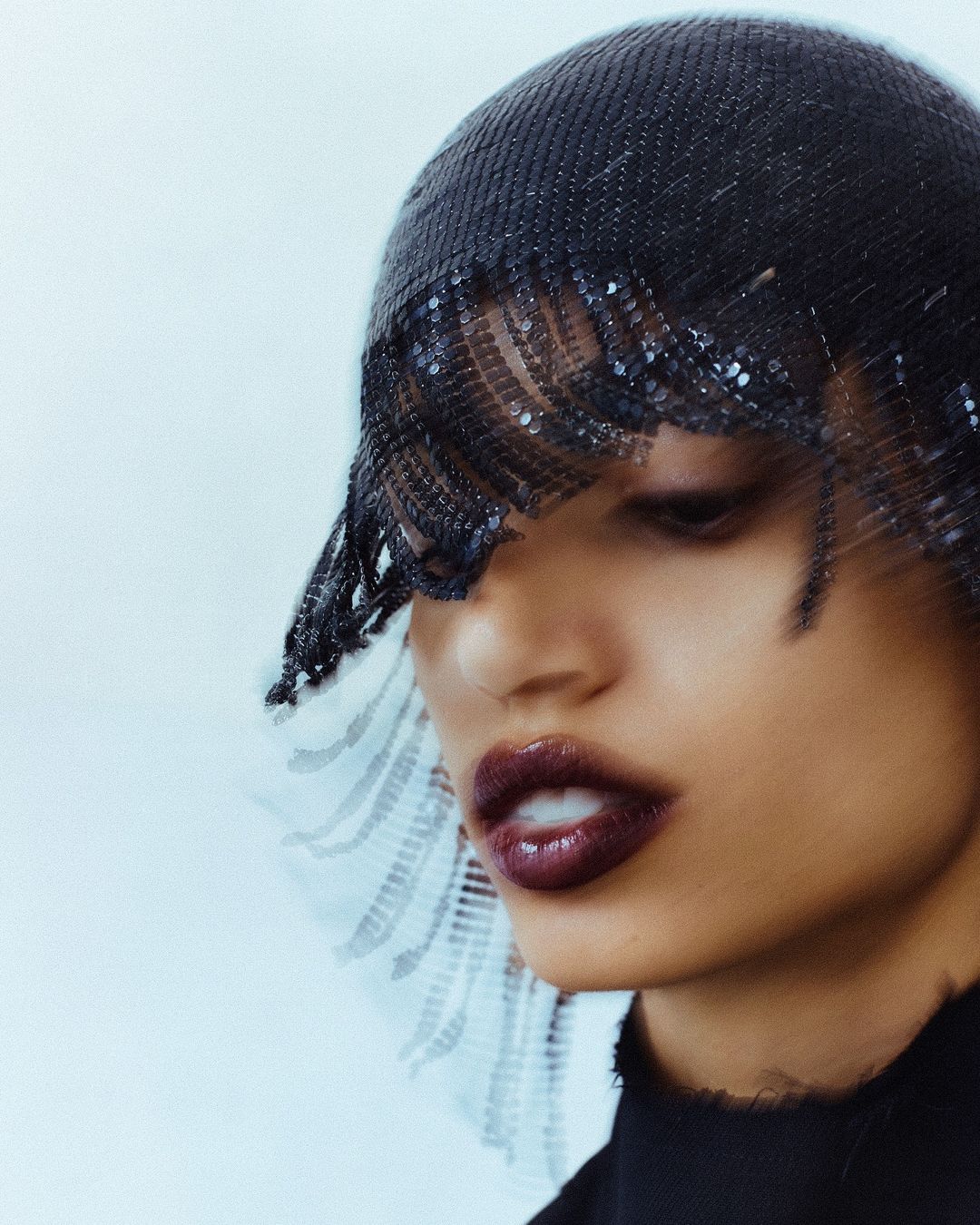 Close-up of a woman with dark hair, wearing a dark, beaded hat with fringe that covers her face, and wearing dark lipstick.