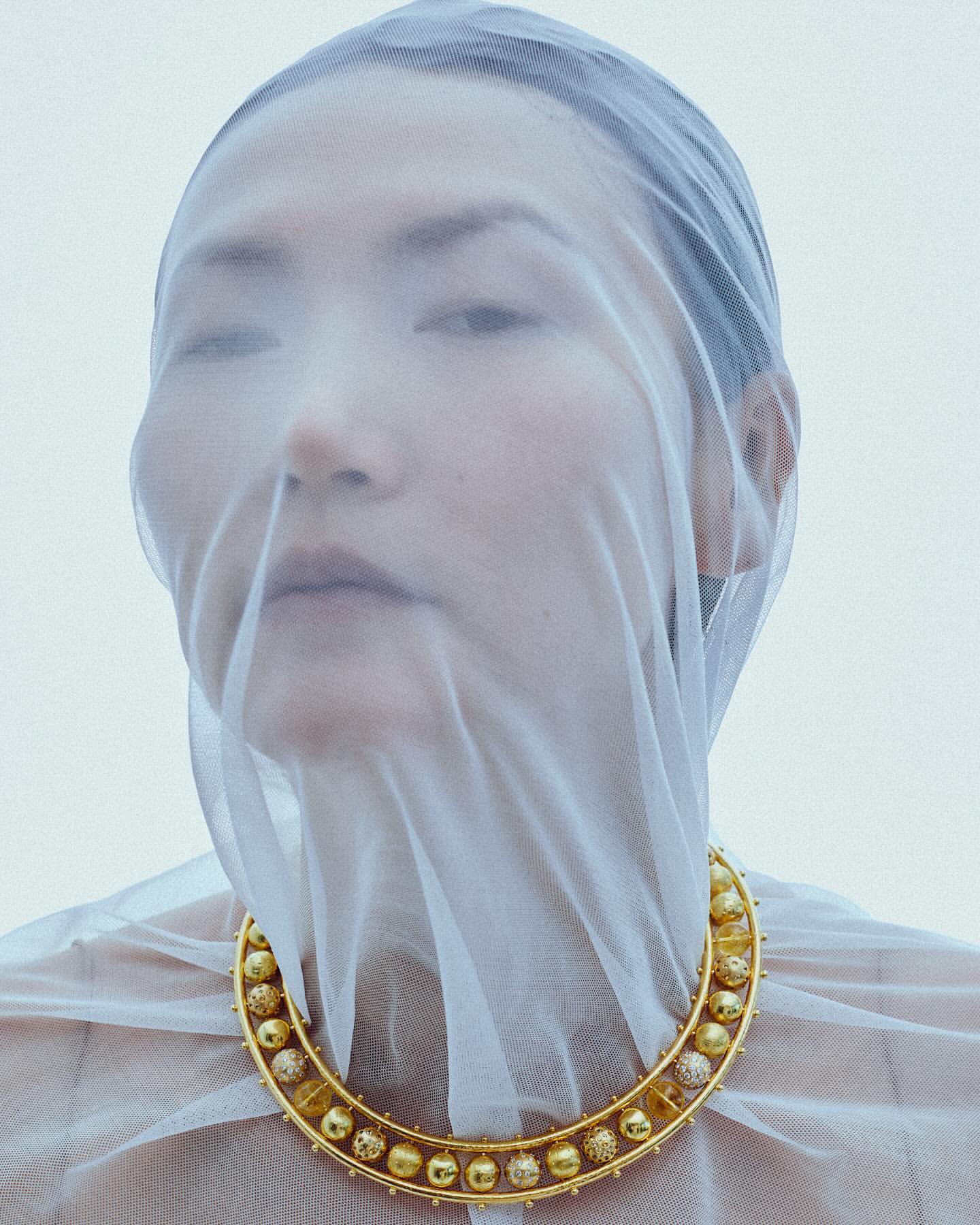 Woman wearing gold beaded necklace and sheer white fabric over her head in a studio shot with a neutral background.