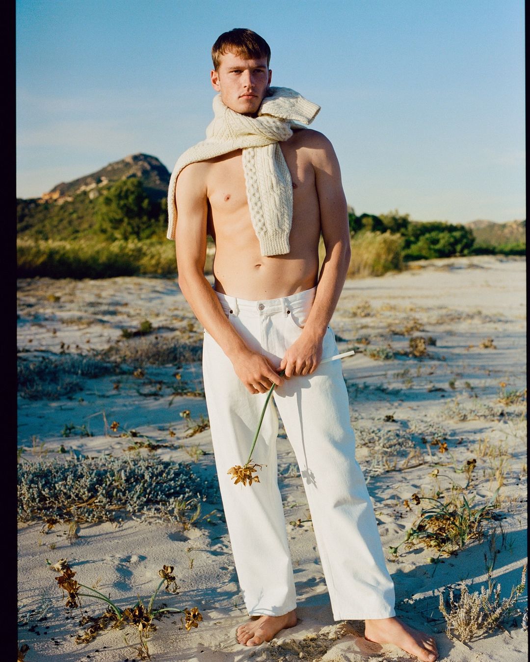 A fair-skinned adult male stands on a beach wearing white pants and a scarf, holding a wilting flower, with a background of dunes and sky.