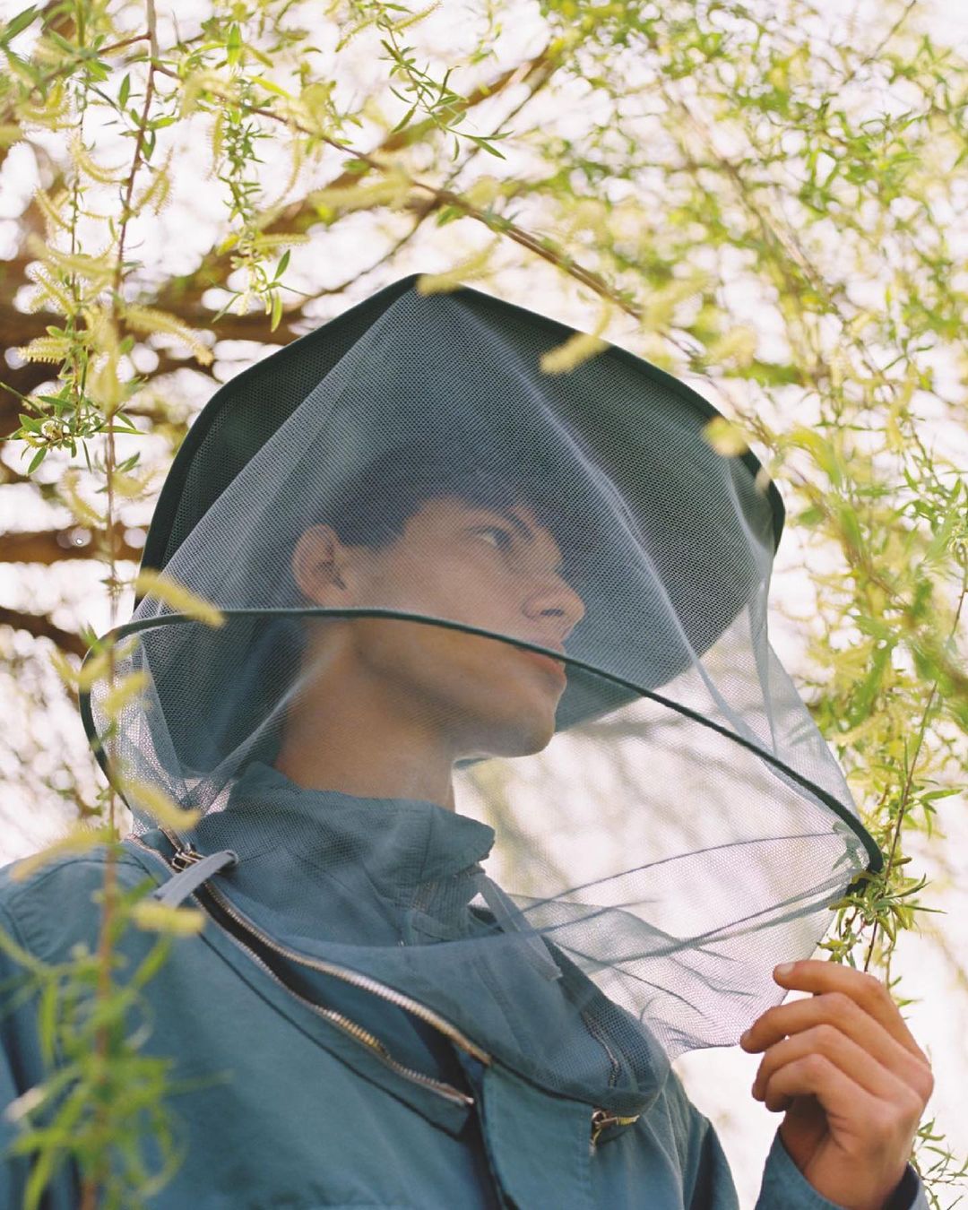 Man in beekeeper veil outdoors surrounded by green foliage. Close-up portrait with shallow depth of field focusing on the person.
