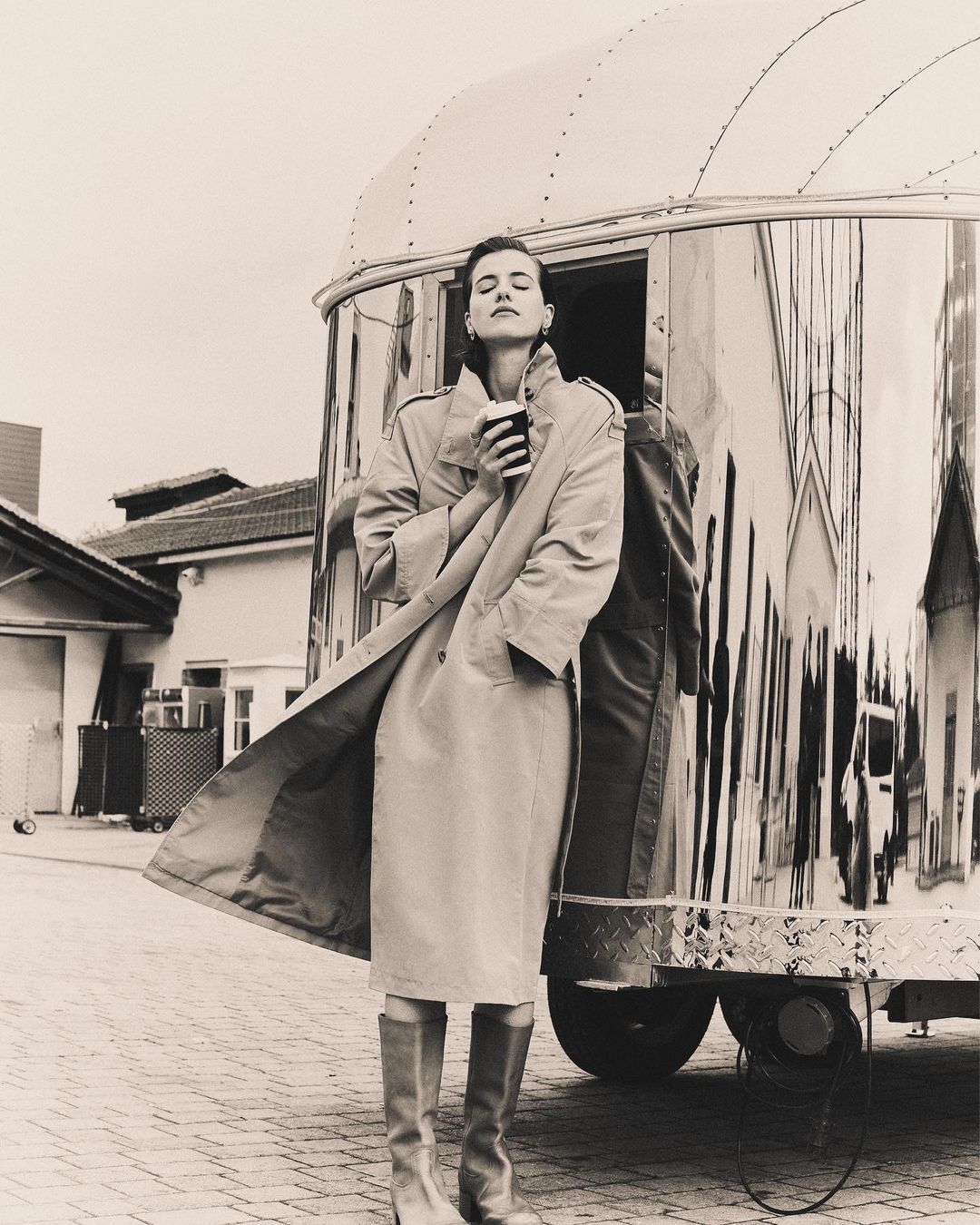 Monochrome fashion shot: Woman in tan coat and boots holds a cup, standing by a shiny trailer on a brick surface.