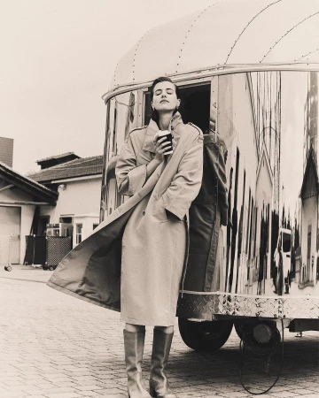 Monochrome fashion shot: Woman in tan coat and boots holds a cup, standing by a shiny trailer on a brick surface.