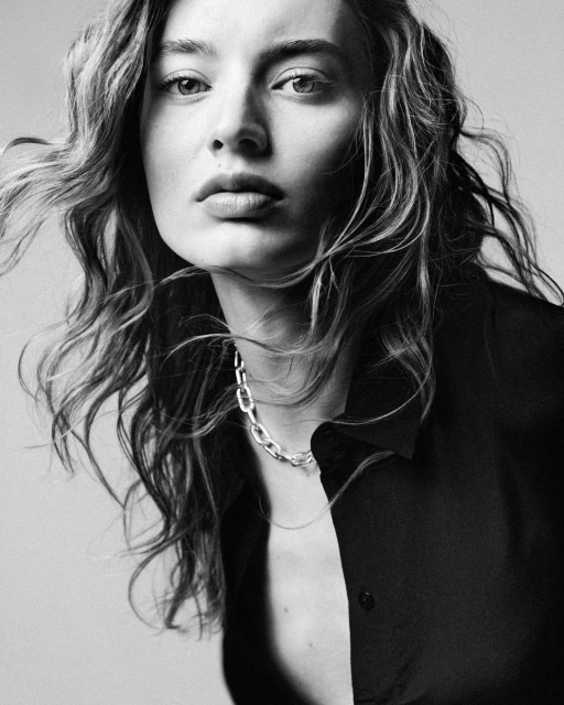 Striking monochrome portrait of a young woman with wavy hair, wearing a chain necklace and unbuttoned dark blouse, gazing intensely.
